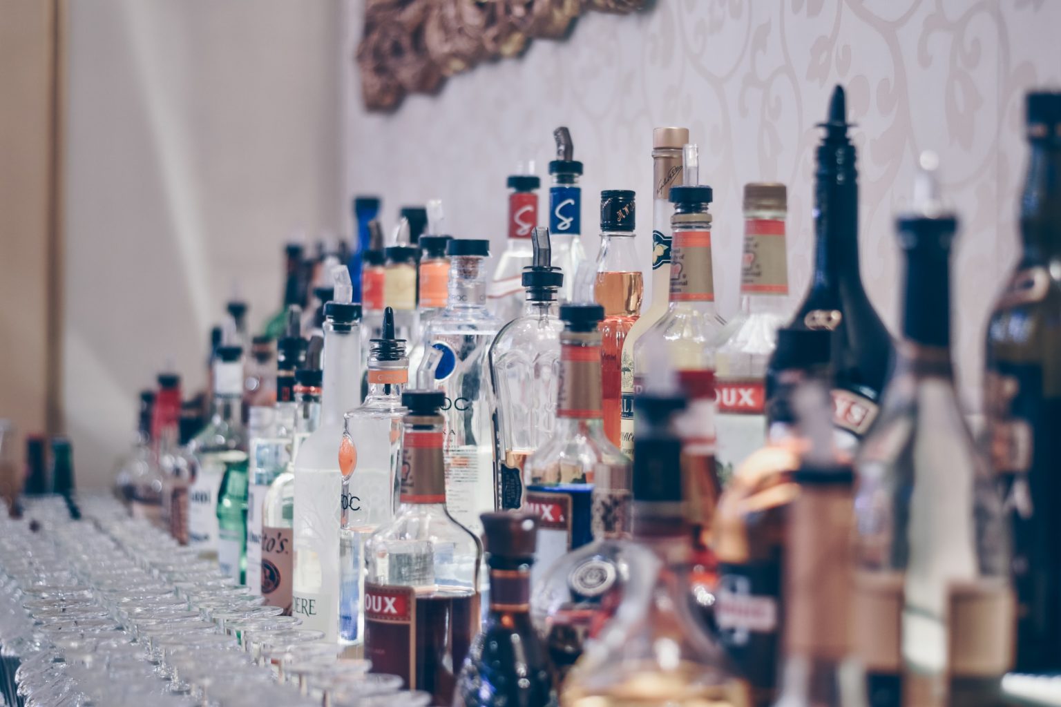 Bottles of alcohol stacked in rows on the back of a bar by a wall. Image by ibrahim-boran on Unsplash.
