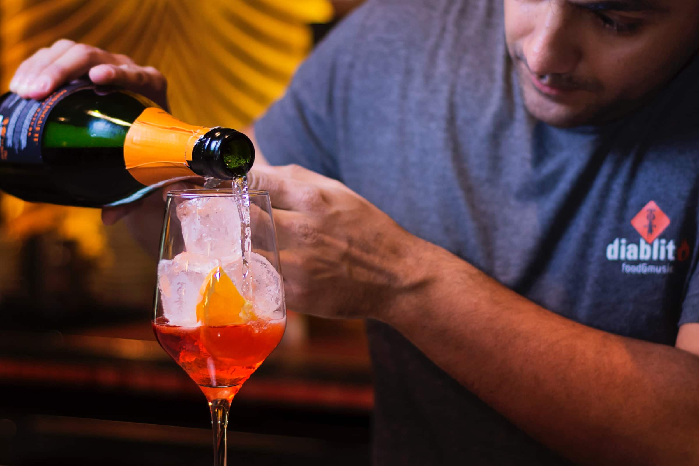 A male bartender shows how to build a cocktail by pouring sparkling wine into a wine glass filled with ice and an orange liqueur. Photo by Kike Salazar N on Unsplash
