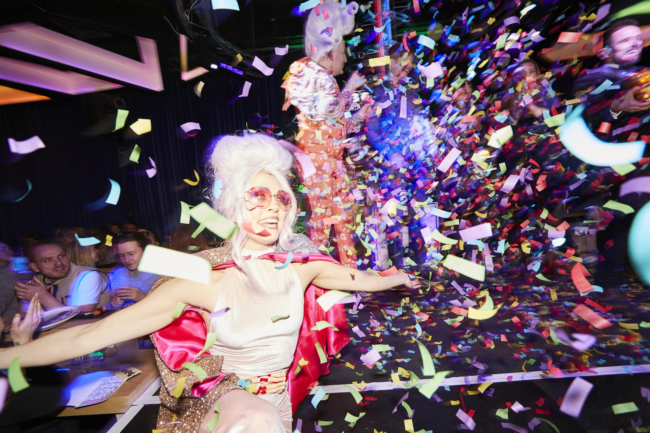 A blonde female entertainer in a glittery costume and bright pink sunglasses dances in as bright confetti falls inside the press launch at Dabblers Bingo, London