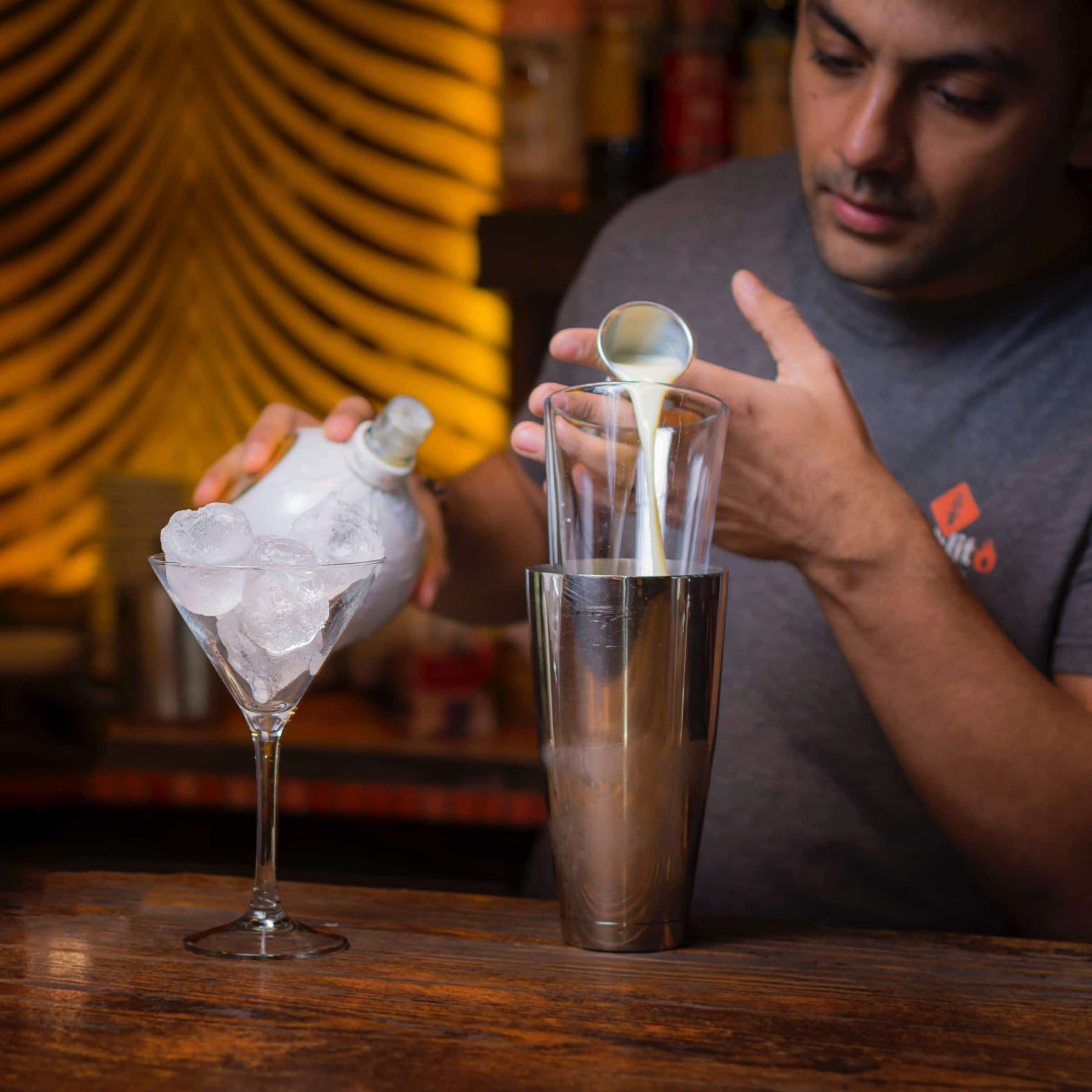 A bartender pours a white liqueur into a Boston Shaker, next to a martini glass filled with ice