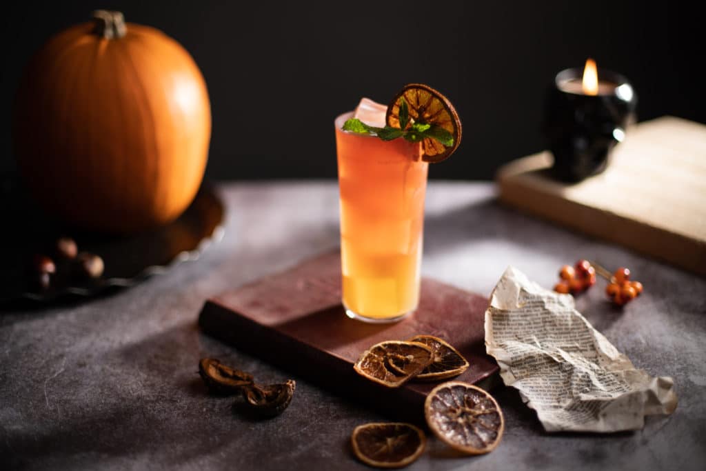The Zombie Halloween cocktail recipe next to a pumpkin, a black Halloween skull and an old book